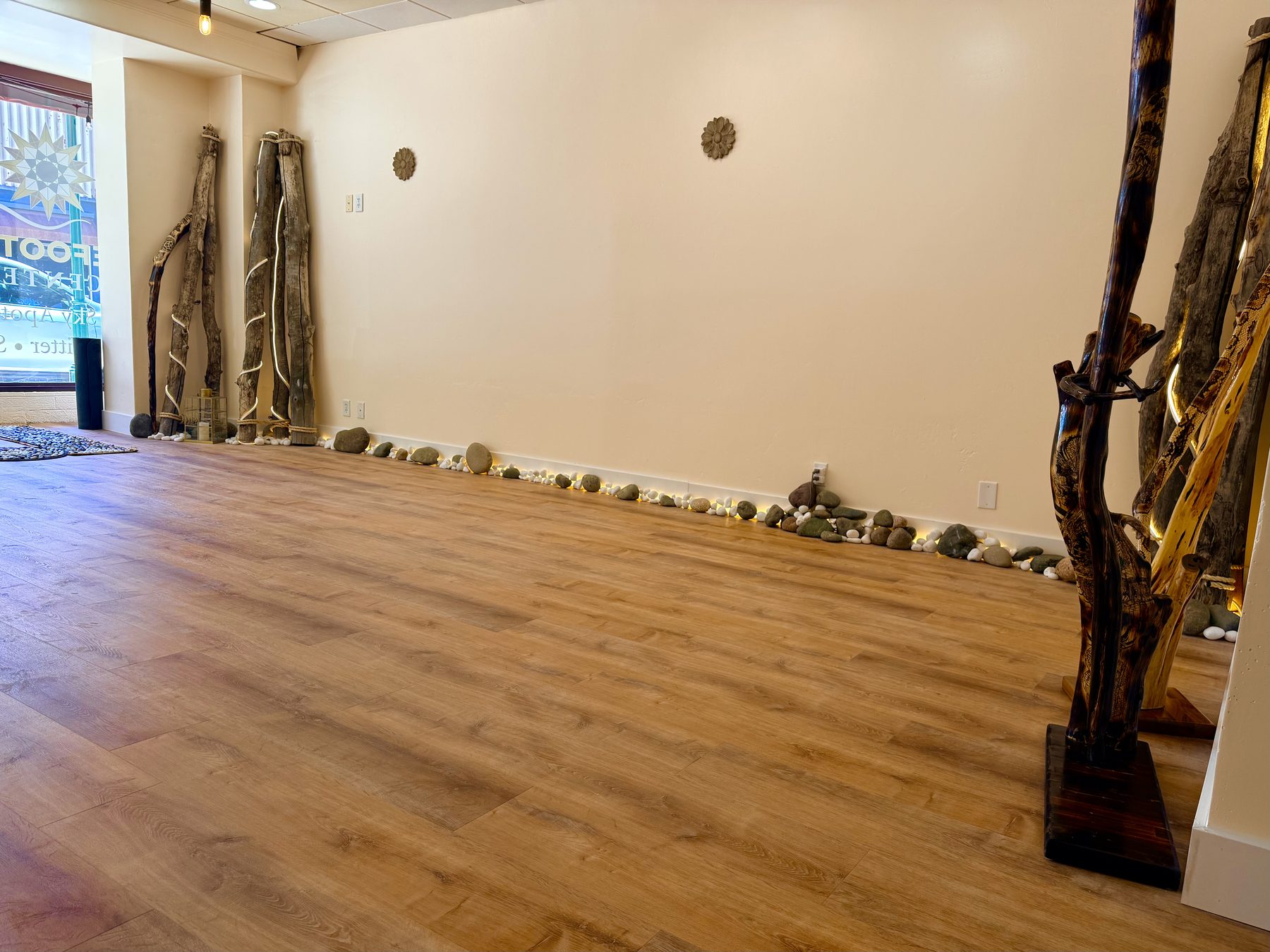The movement classroom at Barefoot Sage Center — bright daylit space with wood floors, driftwood pieces and river stones along the baseboard, the brand mark visible in the window