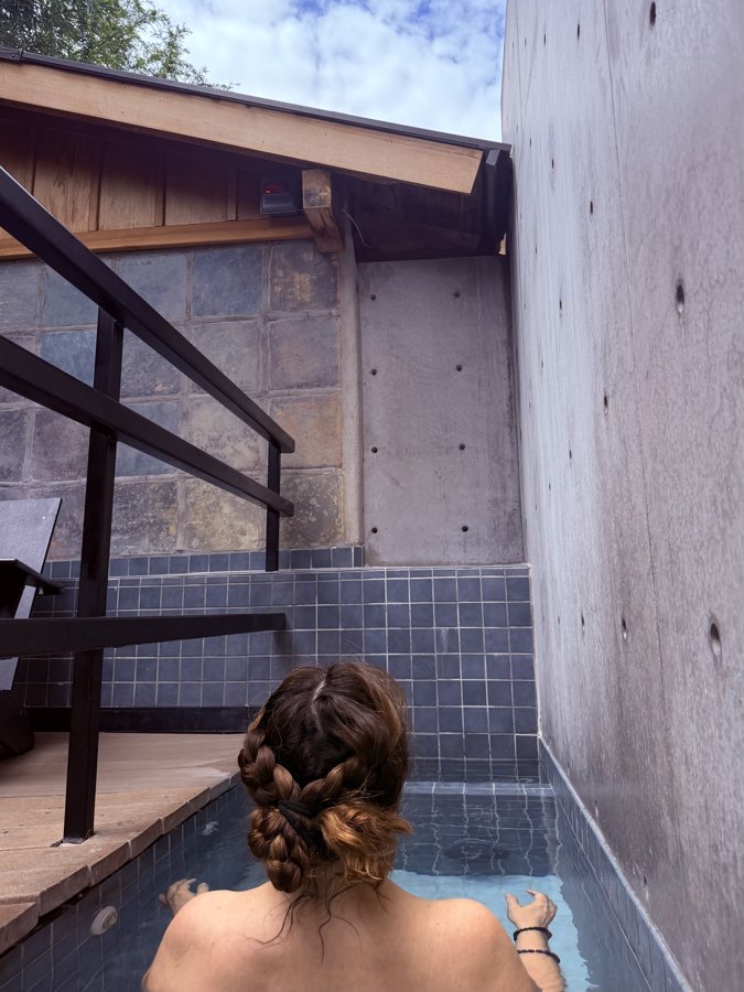 A woman with braided hair sitting in a tiled cold plunge pool, seen from behind