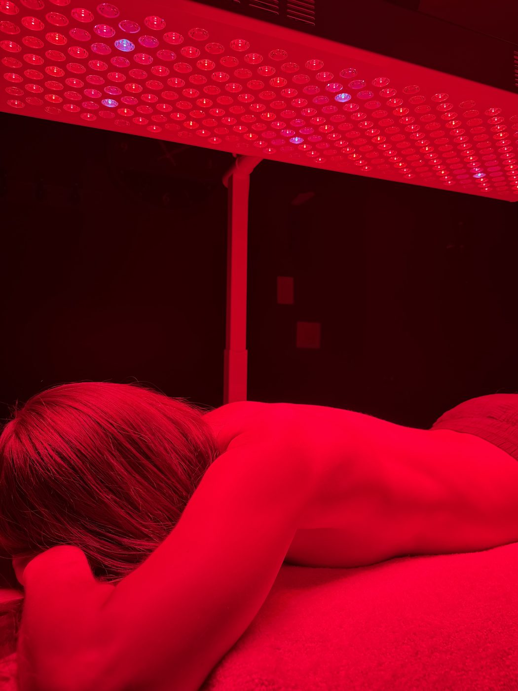 Red-light therapy at Barefoot Sage Center — a guest receiving full-body red and near-infrared light beneath an overhead panel, the room steeped in deep red glow