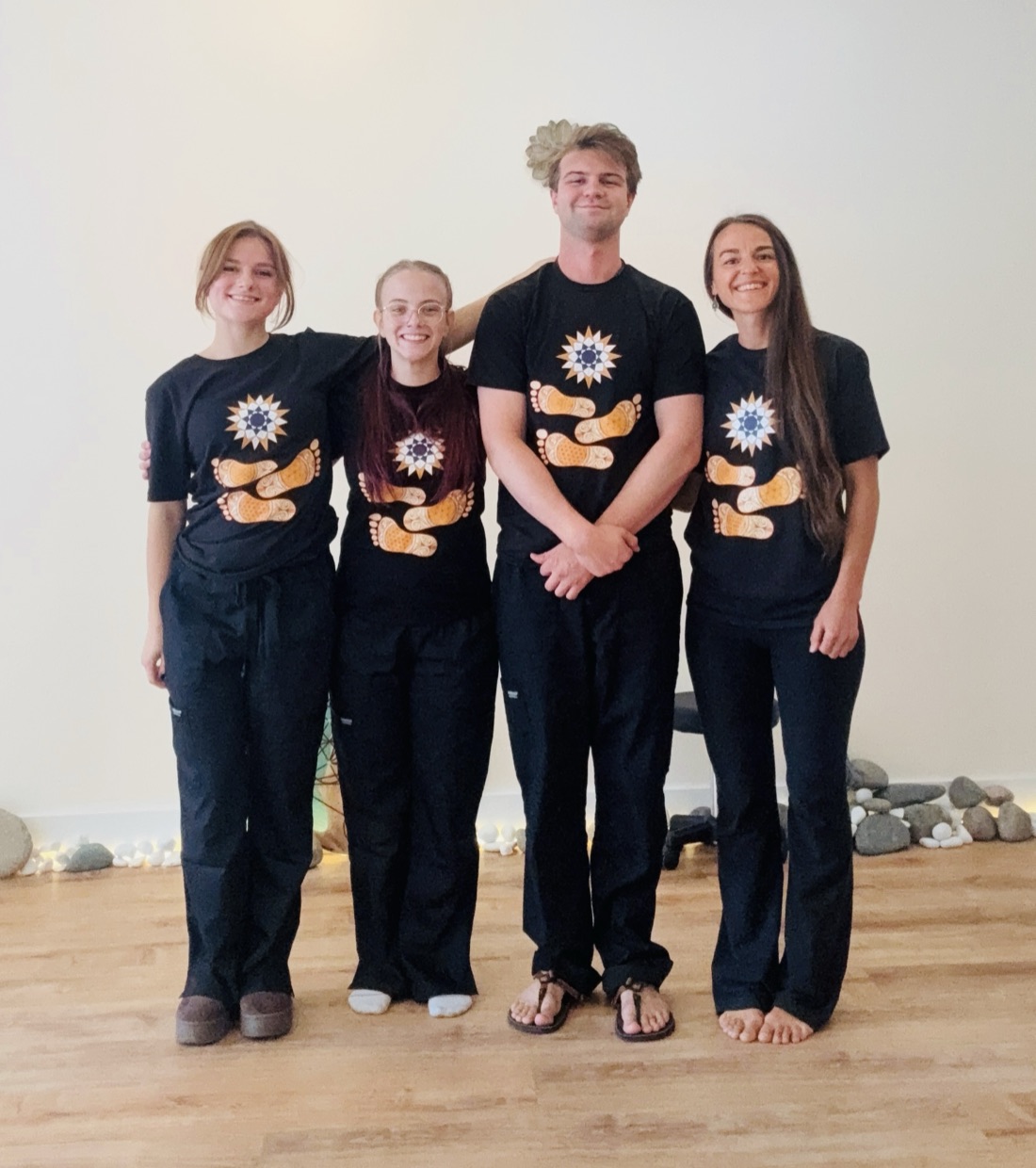 The Barefoot Sage Center practitioners standing together in the studio, wearing black Barefoot Sage shirts, with river stones and warm light along the wall behind them