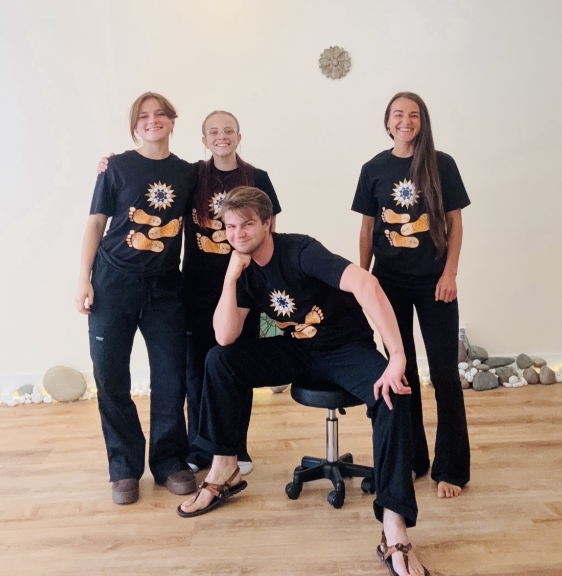 The Barefoot Sage Center team together in the studio — three practitioners standing and one seated on a stool, all in black Barefoot Sage shirts, with river stones and warm light along the wall