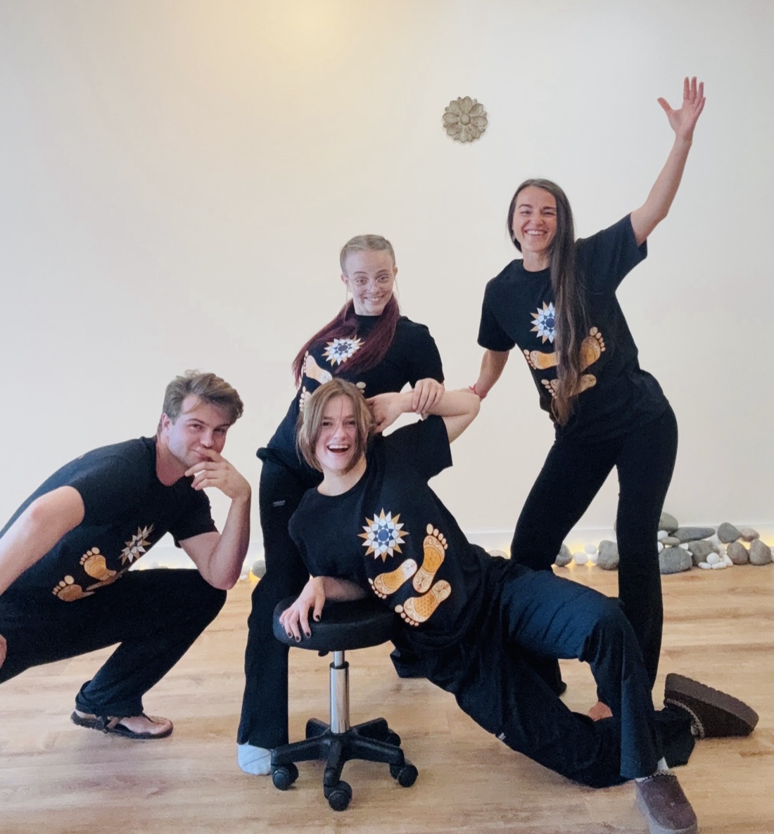 The Barefoot Sage Center team laughing together in the studio — one arm raised, everyone in black Barefoot Sage shirts, on the wooden floor beside the river stones